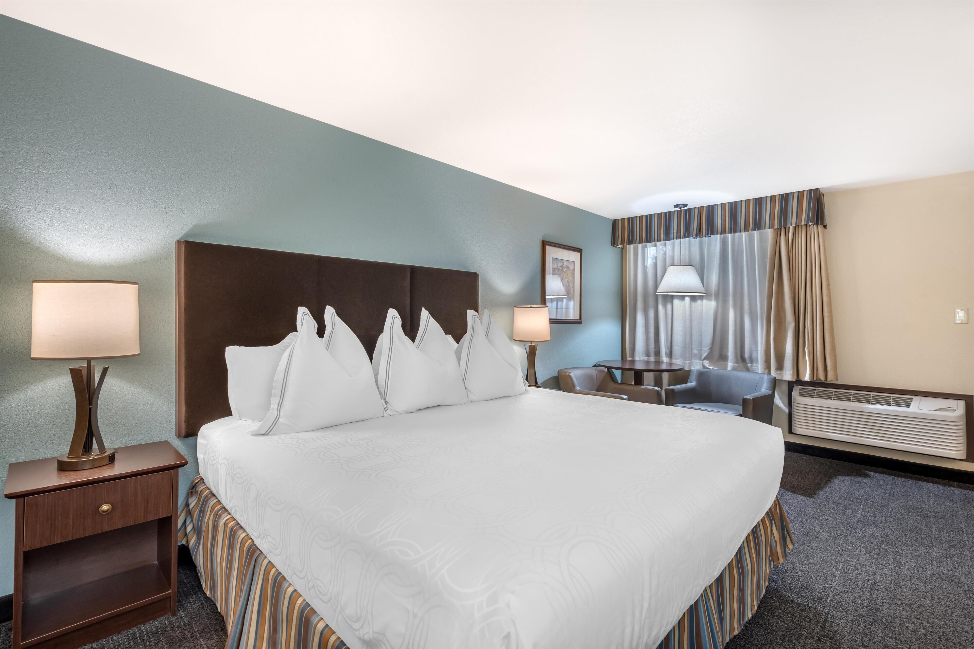 A hotel room with a large bed, two bedside tables with lamps, a desk with a chair, a window with curtains, and a wall-mounted air conditioner.