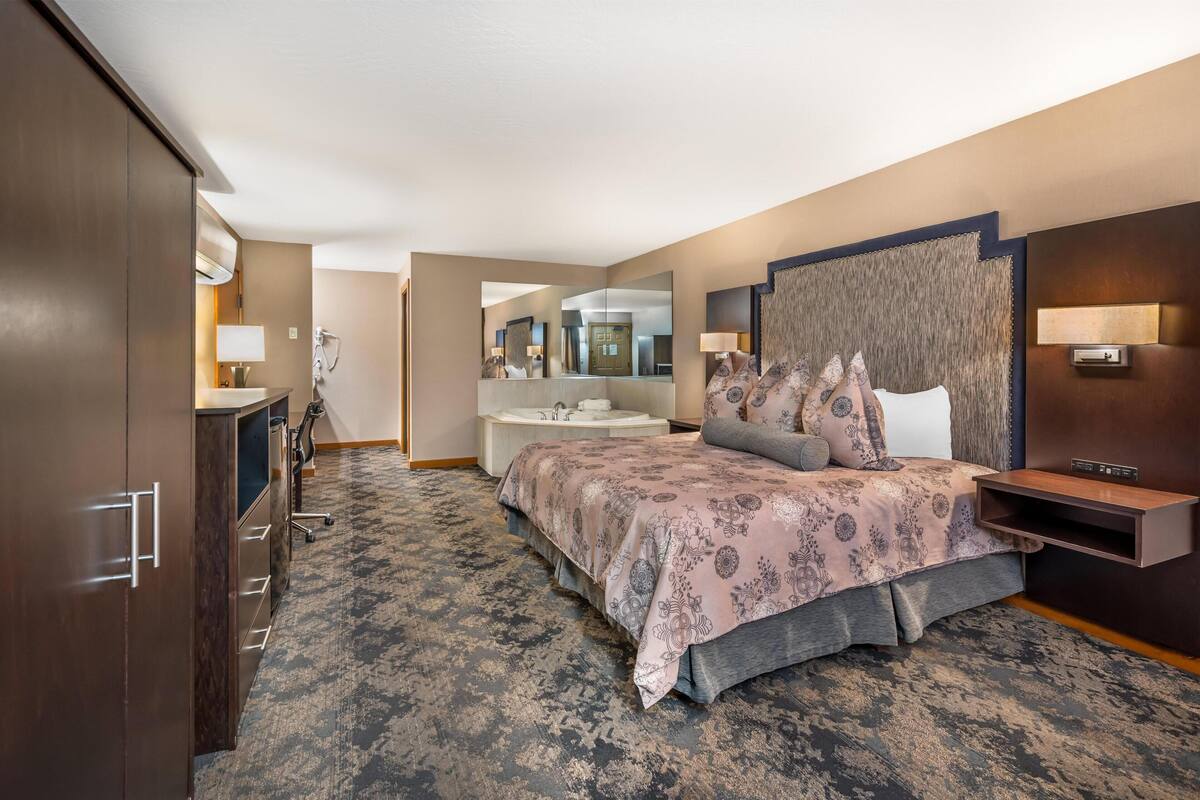 A hotel room with a large bed, a desk, a vanity with a mirror, and a bathroom visible in the background.