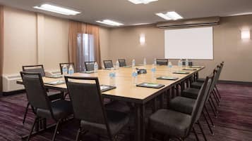 Un salón de conferencias con una larga mesa, sillas, botellas de agua y una pantalla para proyector.