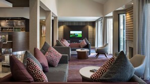 A modern hotel lobby with a grey sofa, armchairs, a coffee table, a TV screen, and a bar area with a menu board.