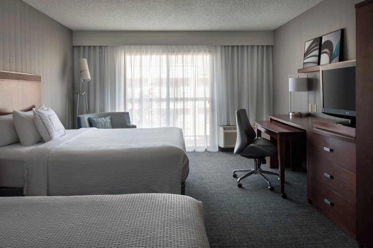 A hotel room with two beds, a desk with a computer, a TV, and a large window with curtains.
