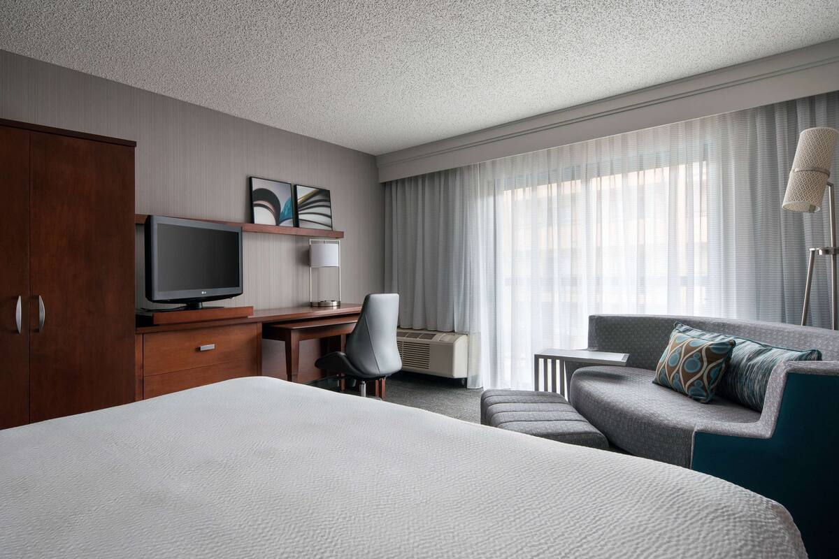 A hotel room with a large bed, a desk with a television, a chair, a sofa, and a large window with curtains.