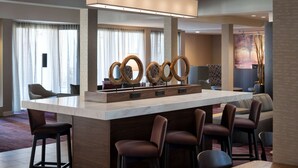 A modern hotel lobby with a marble bar, leather bar stools, and a large decorative mirror.