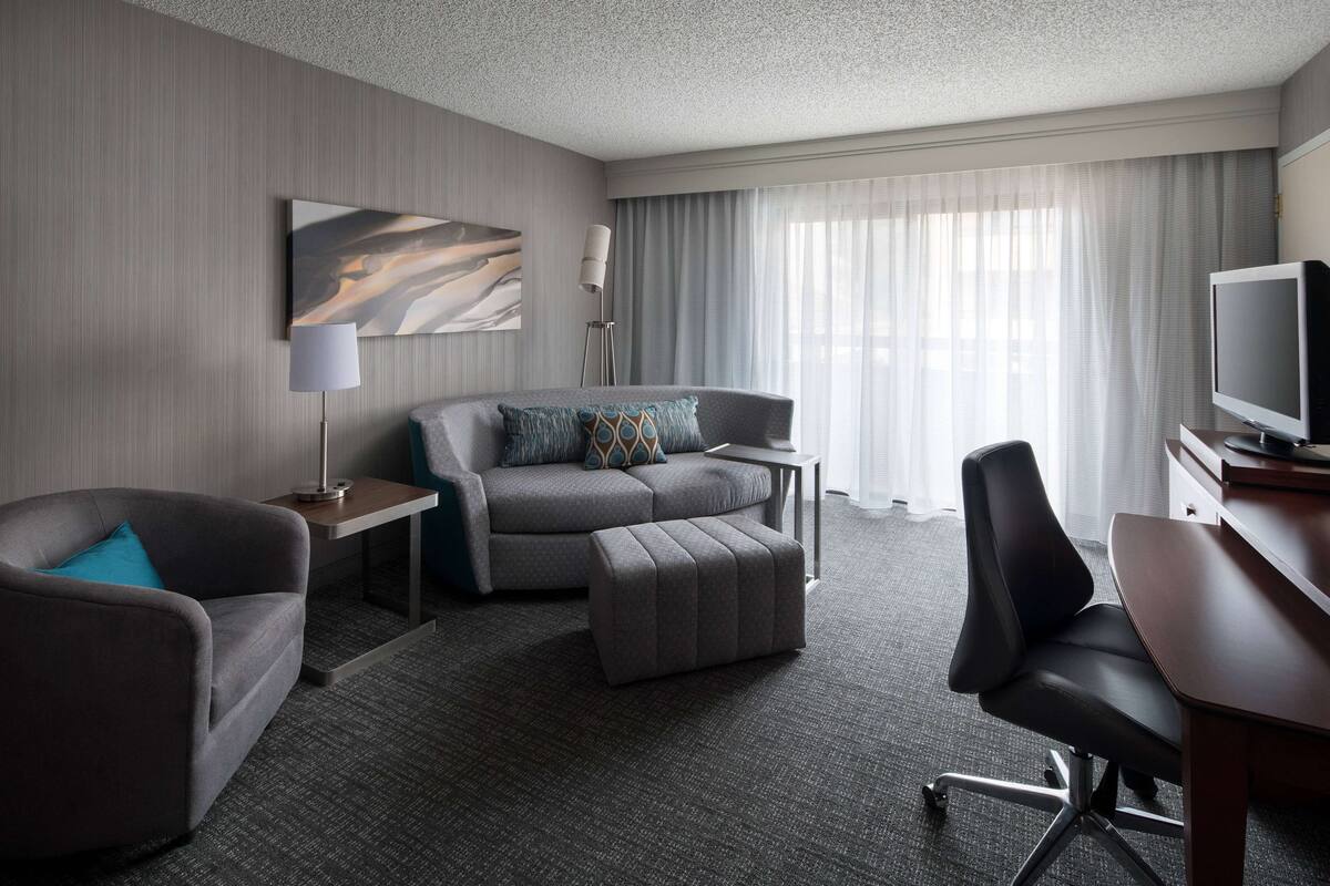 A modern hotel room with a grey sofa, a matching ottoman, a black office chair, a flat-screen TV, a small table with a lamp, and a large window with sheer curtains.