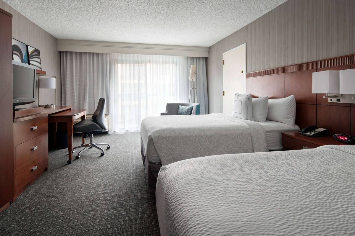 A hotel room with two beds, a desk with a chair, a TV, and a lamp.