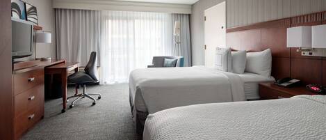 A hotel room with two beds, a desk with a chair, a TV, and a lamp.