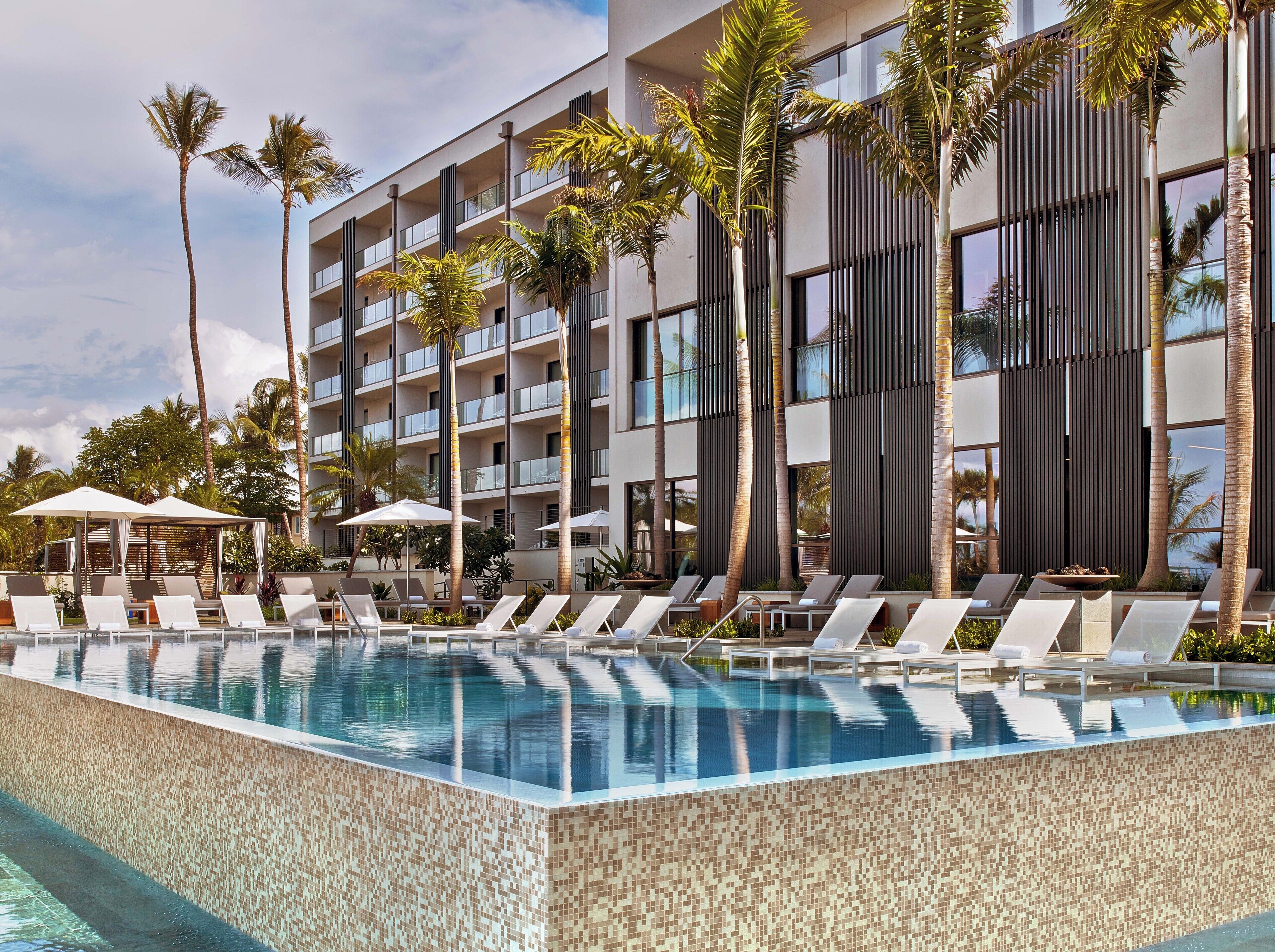 A modern hotel with a pool area, lounge chairs, and palm trees.