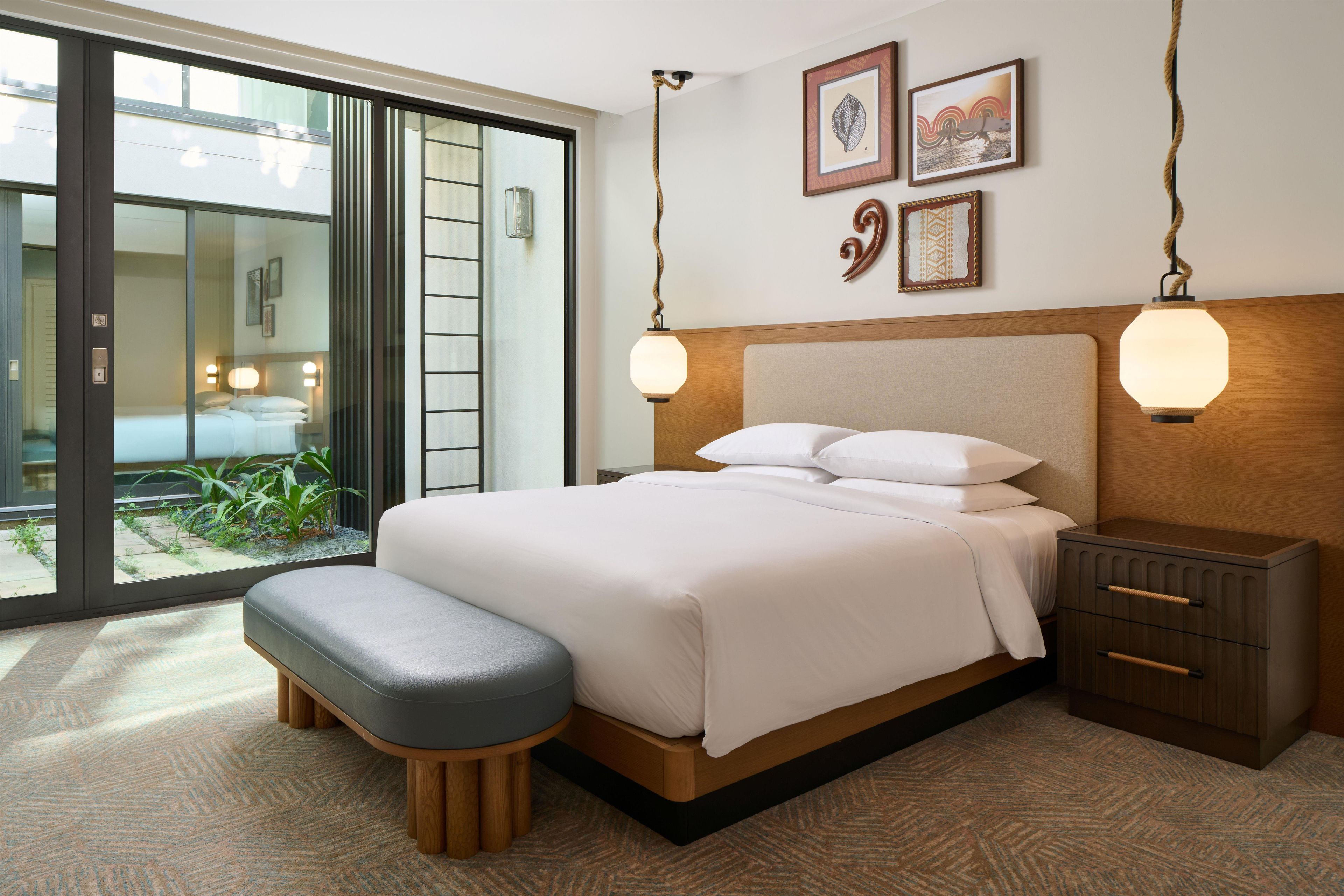 A hotel room with a bed, bedside tables, a bench, a view of a garden through a sliding door, and artwork on the wall.