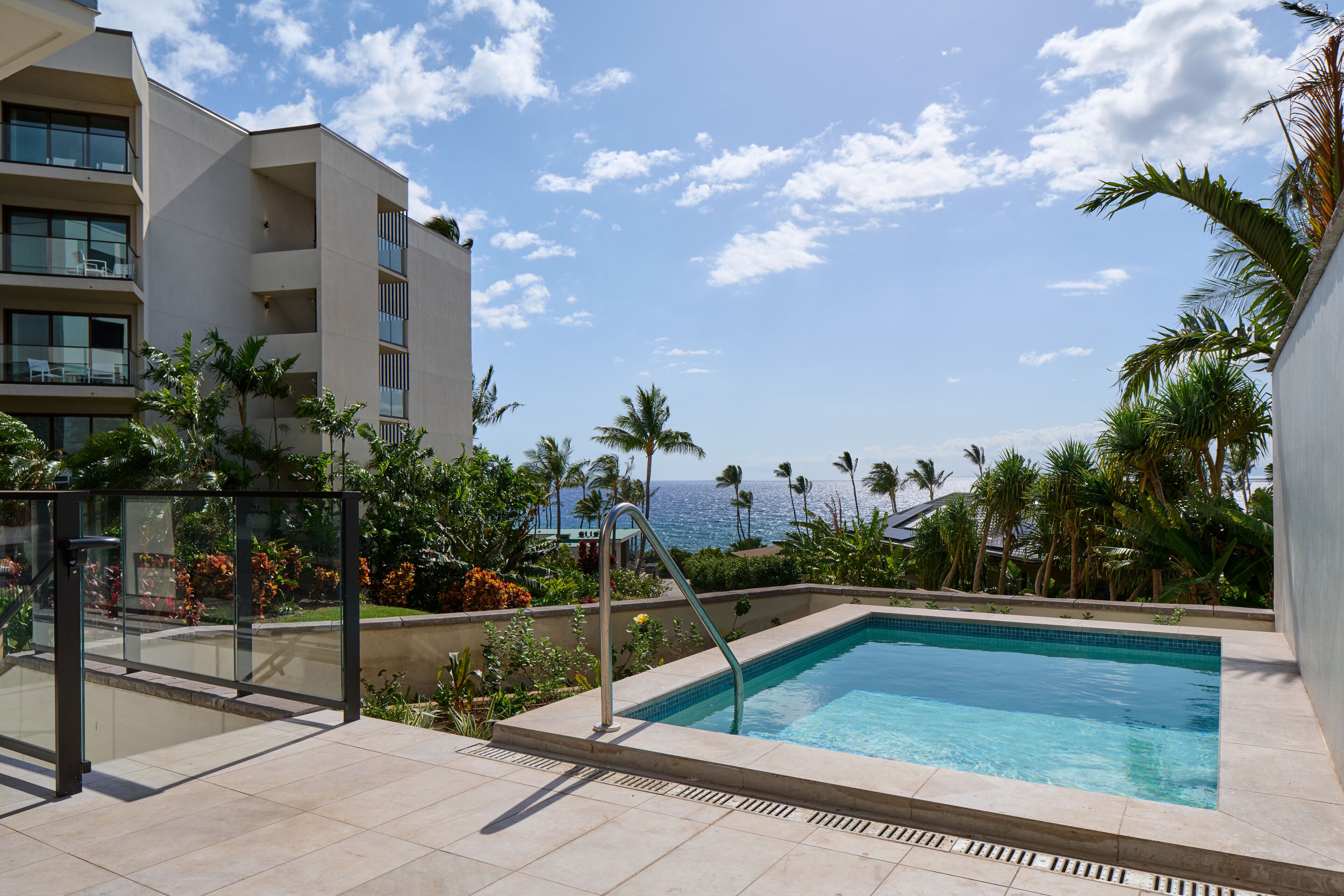 Zona de piscina con vista despejada al océano, rodeada de palmeras y un edificio con balcones.