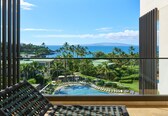 A balcony with a view of a pool, palm trees, and a distant coastline.