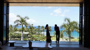 A room with a large glass door leading to a balcony overlooking a tropical landscape with palm trees and a view of the ocean.