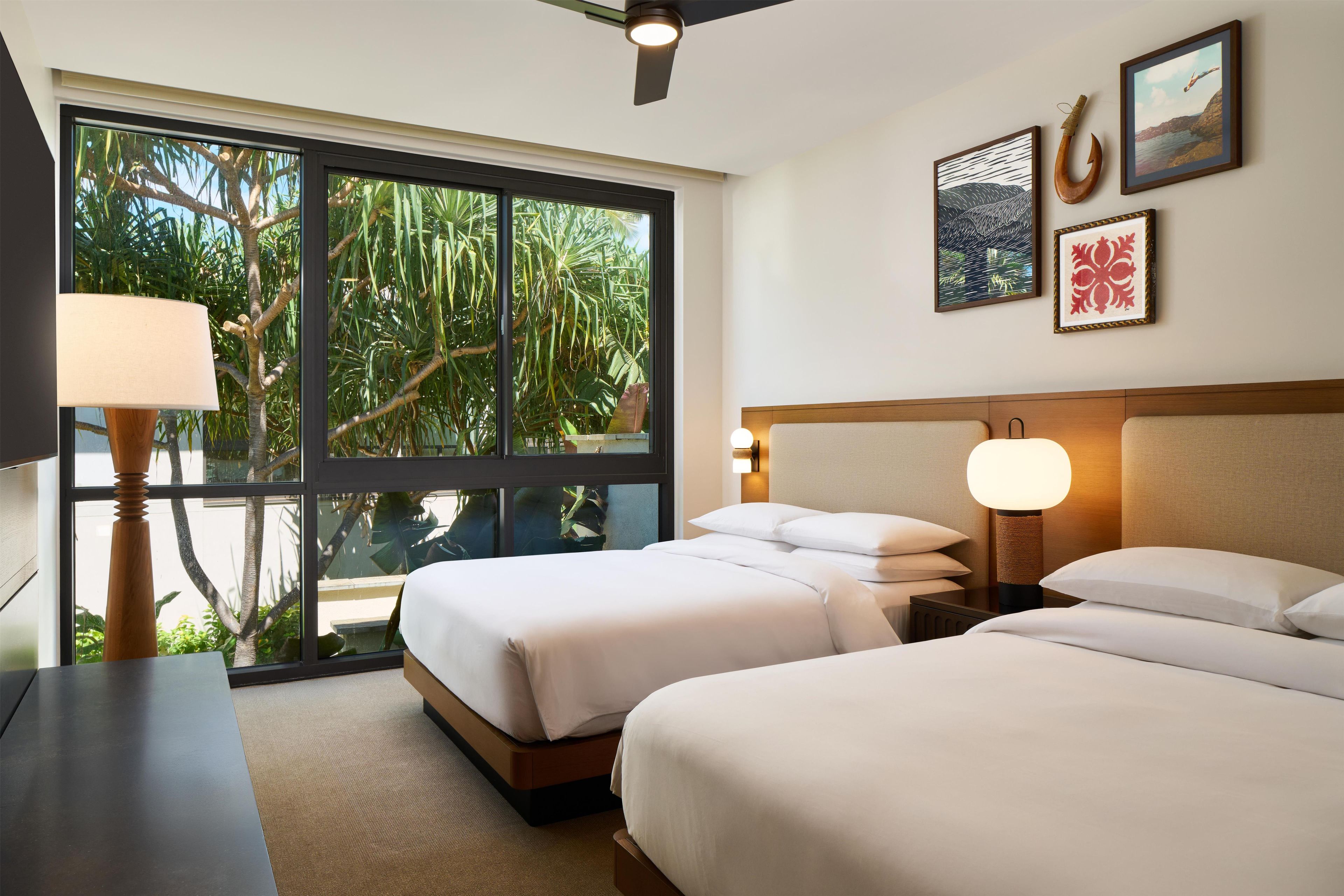 A hotel room with two beds, a ceiling fan, a large window with a view of greenery, and wall-mounted artwork.