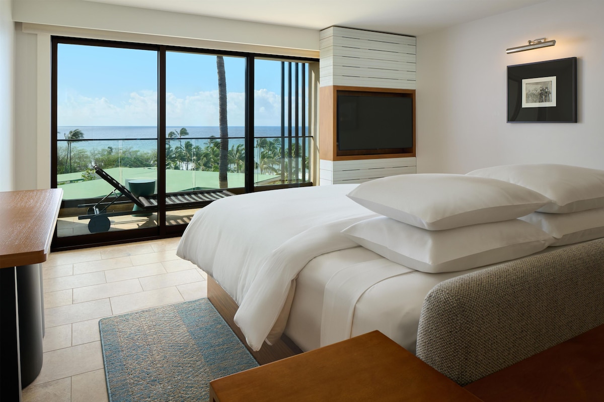 Een slaapkamer met een groot raam, een bed met witte lakens, een televisie en uitzicht op de oceaan.