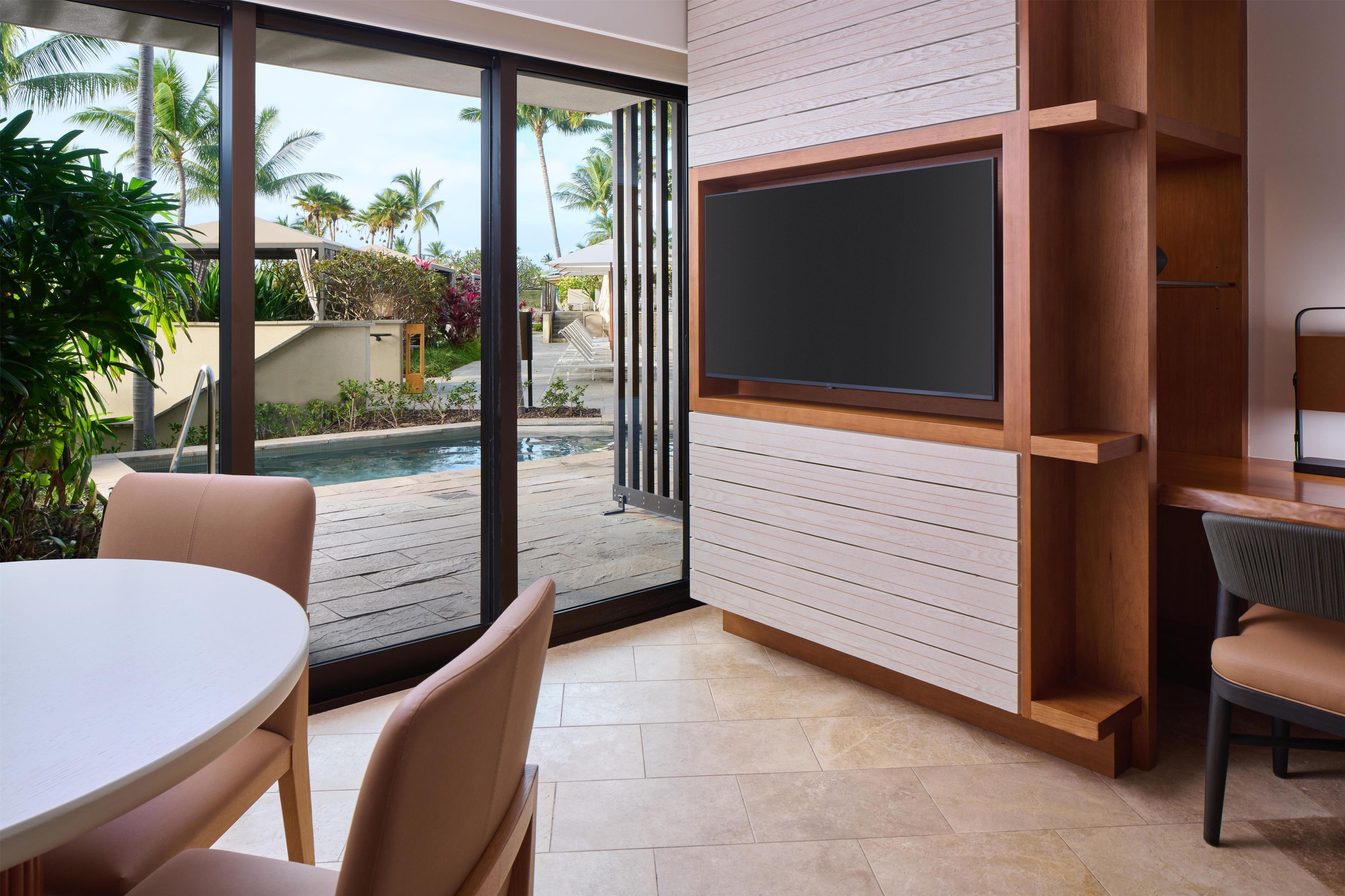 Una sala de estar moderna con un televisor grande montado en una pared de madera, una piscina visible a través de la puerta de cristal, y un área de descanso con una mesa redonda y sillas.