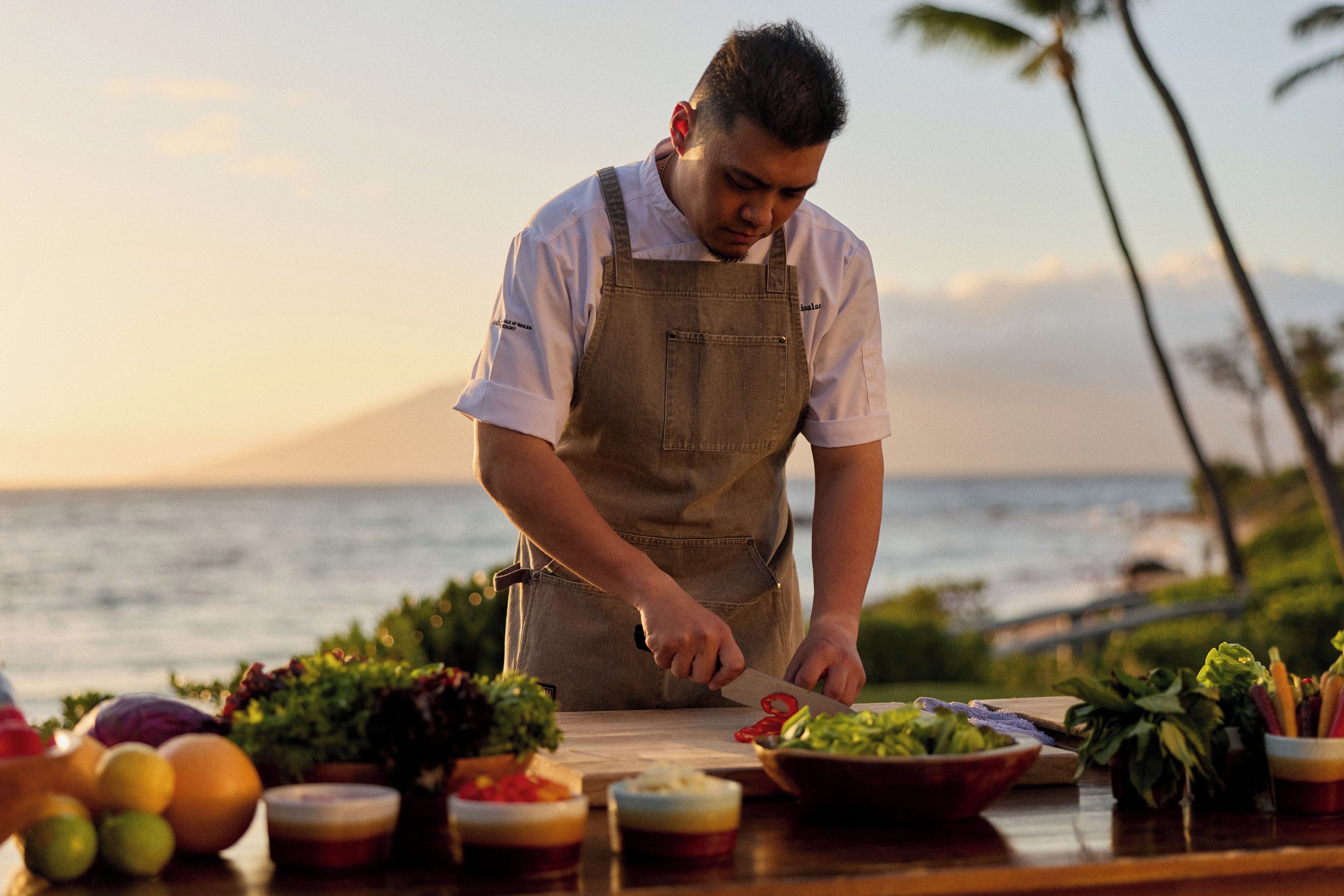 A chef preparing food at a wooden table with various ingredients and bowls, overlooking a body of water.