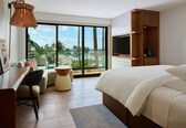 A hotel room with a bed, a desk, a chair, a TV, and a view of palm trees and the ocean.