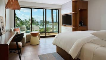 Habitación de hotel con cama, escritorio, silla, televisor y vista a palmeras y al océano.