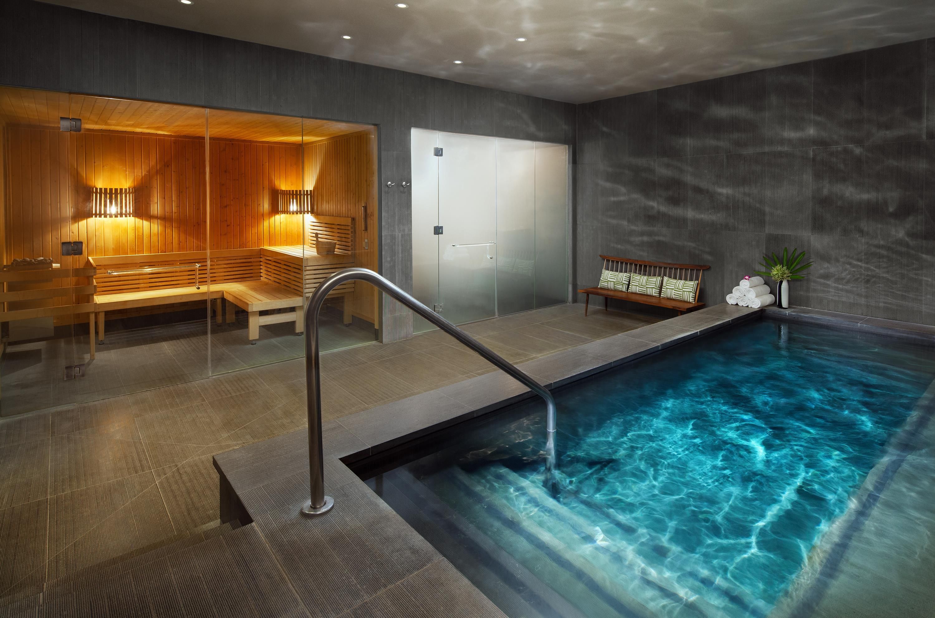 A modern indoor swimming pool with a wooden bench and a glass door leading to a sauna area.