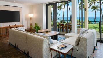 A modern living room with a large flat-screen TV, a sofa set, a coffee table, and a dining area with a view of the ocean.