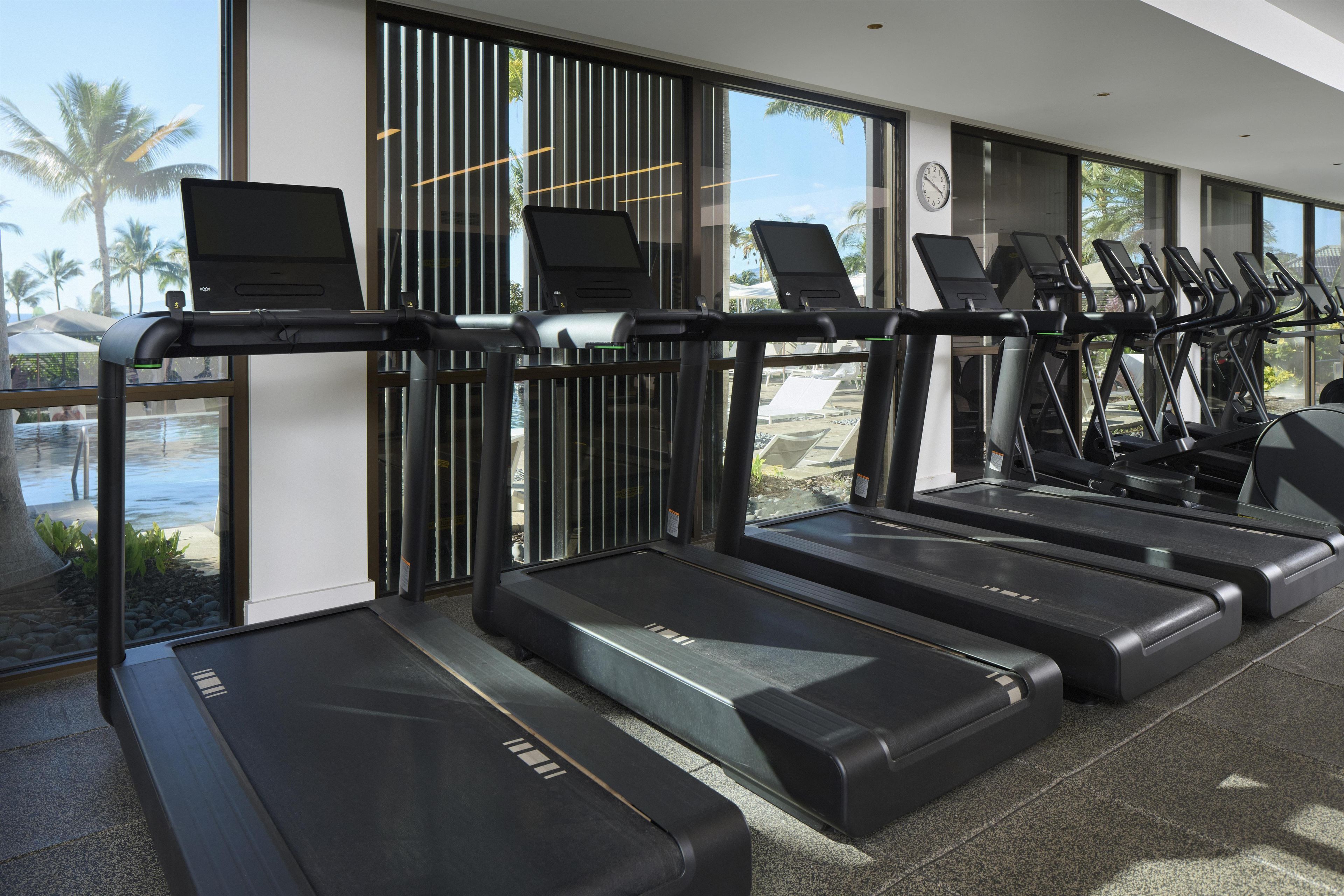 A gym with multiple treadmills, a pool visible through the windows, and a clock on the wall.