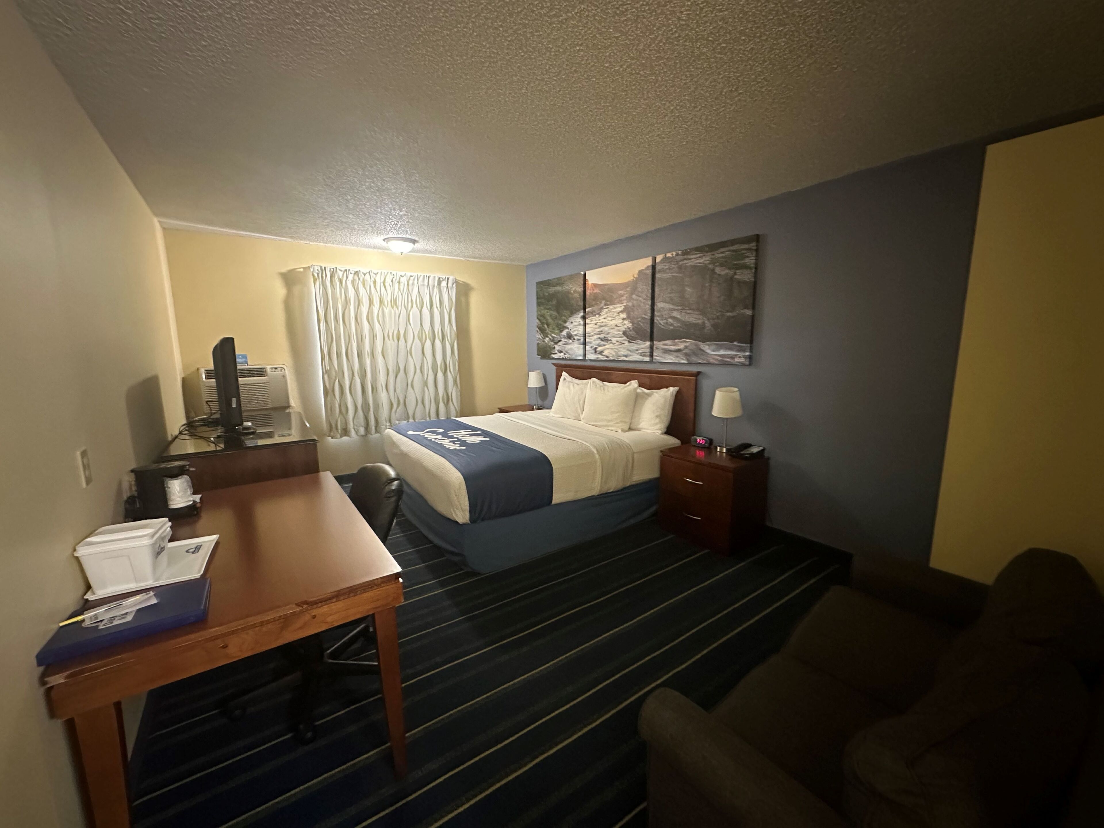 A hotel room with a bed, a desk, a chair, a couch, a painting on the wall, and a window with curtains.