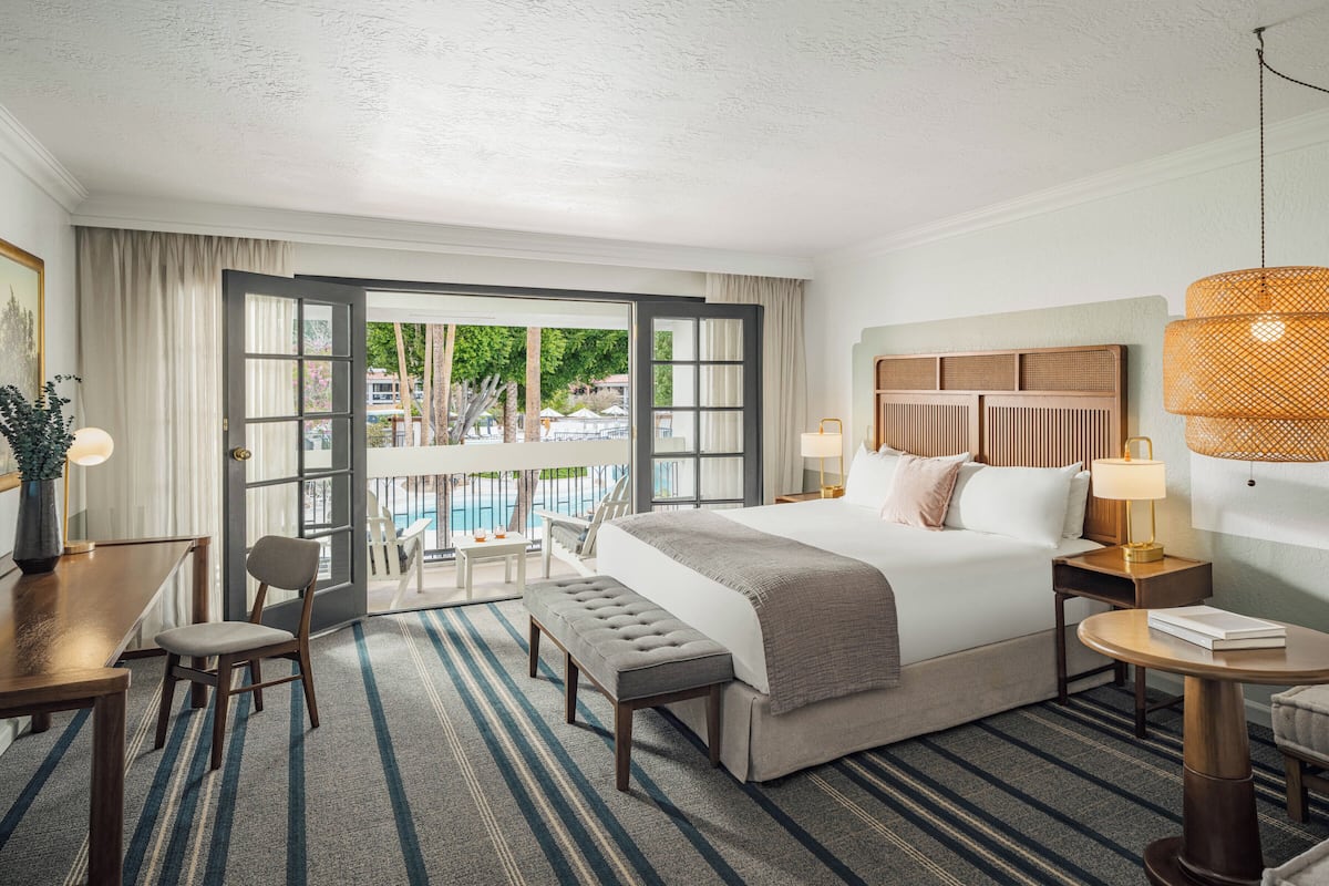 A hotel room with a bed, bedside tables, a desk, a chair, a bench, a large window with a view of a pool and trees, and a ceiling light fixture.