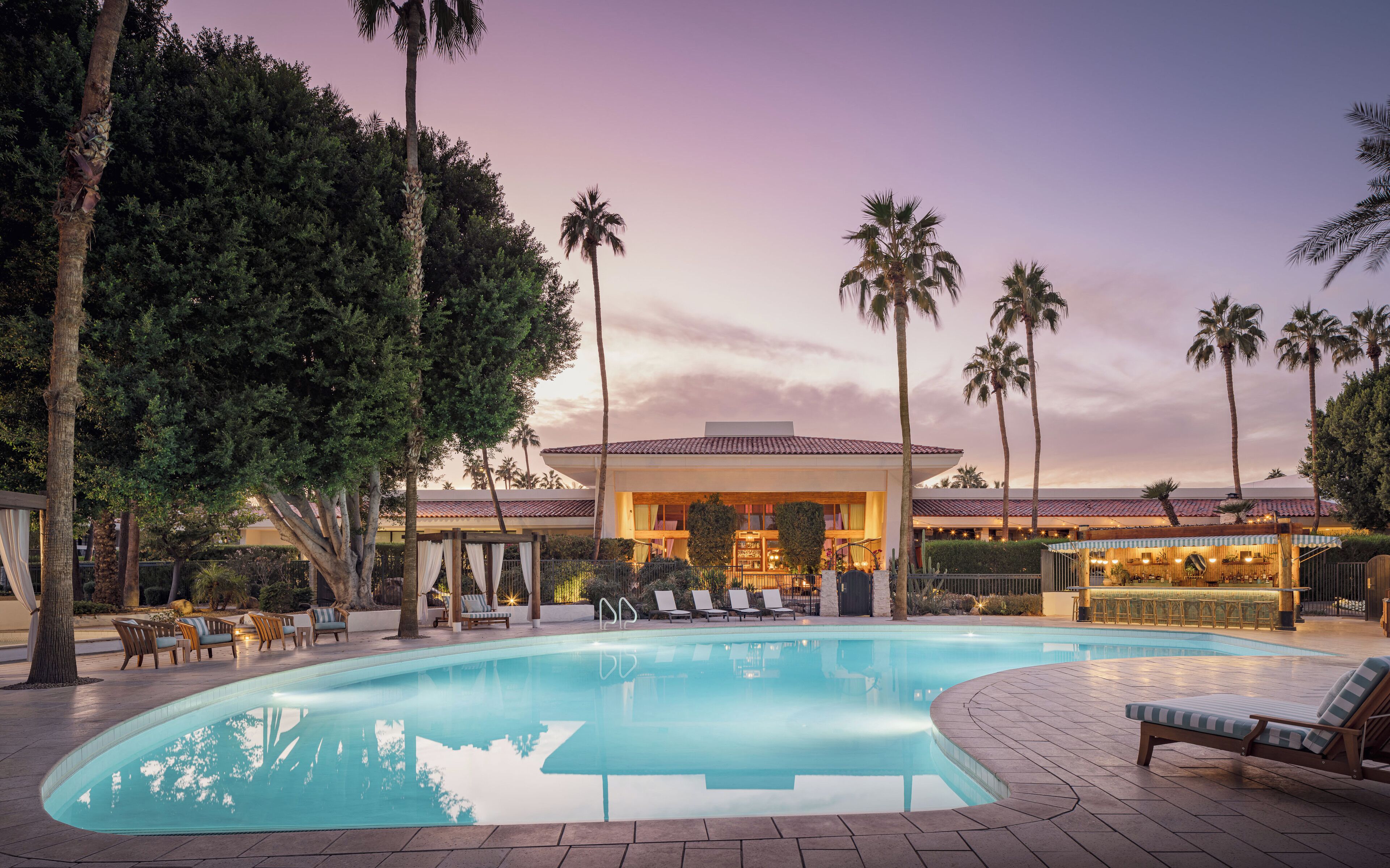 A pool area with lounge chairs and a bar, surrounded by palm trees and a building with a tiled roof.