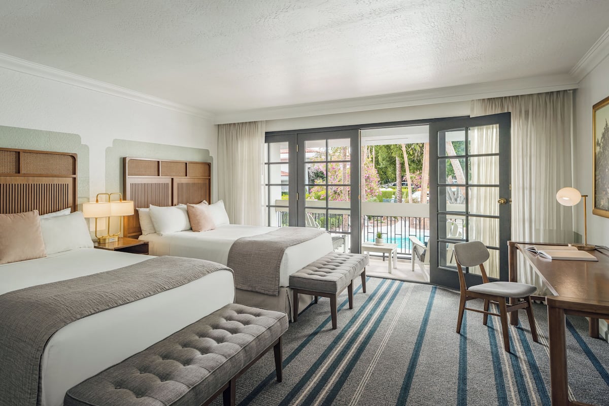 A hotel room with two beds, a desk, a chair, a bench, and a view of the outdoor area.