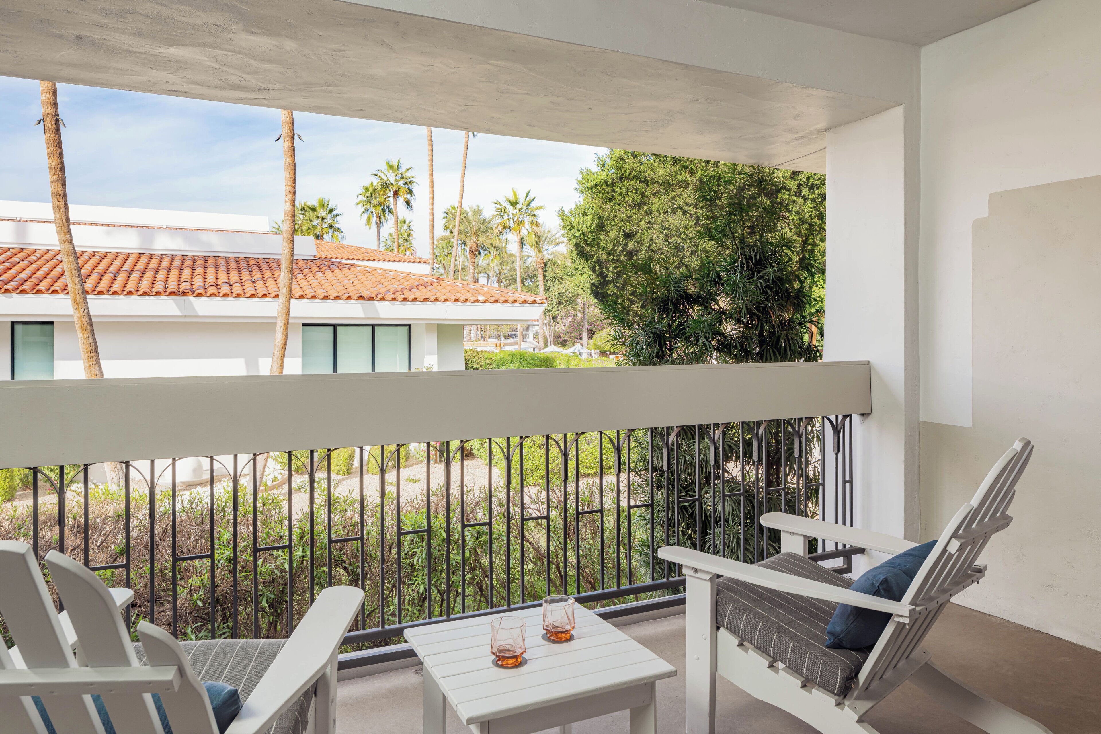 A balcony with white chairs, a small table, and a view of a building with a tiled roof and palm trees.