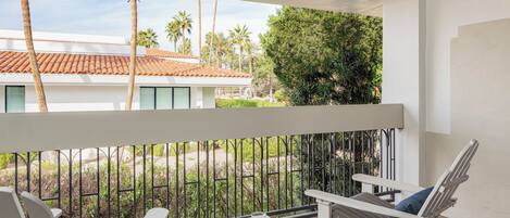 A balcony with white chairs, a small table, and a view of a building with a tiled roof and palm trees.