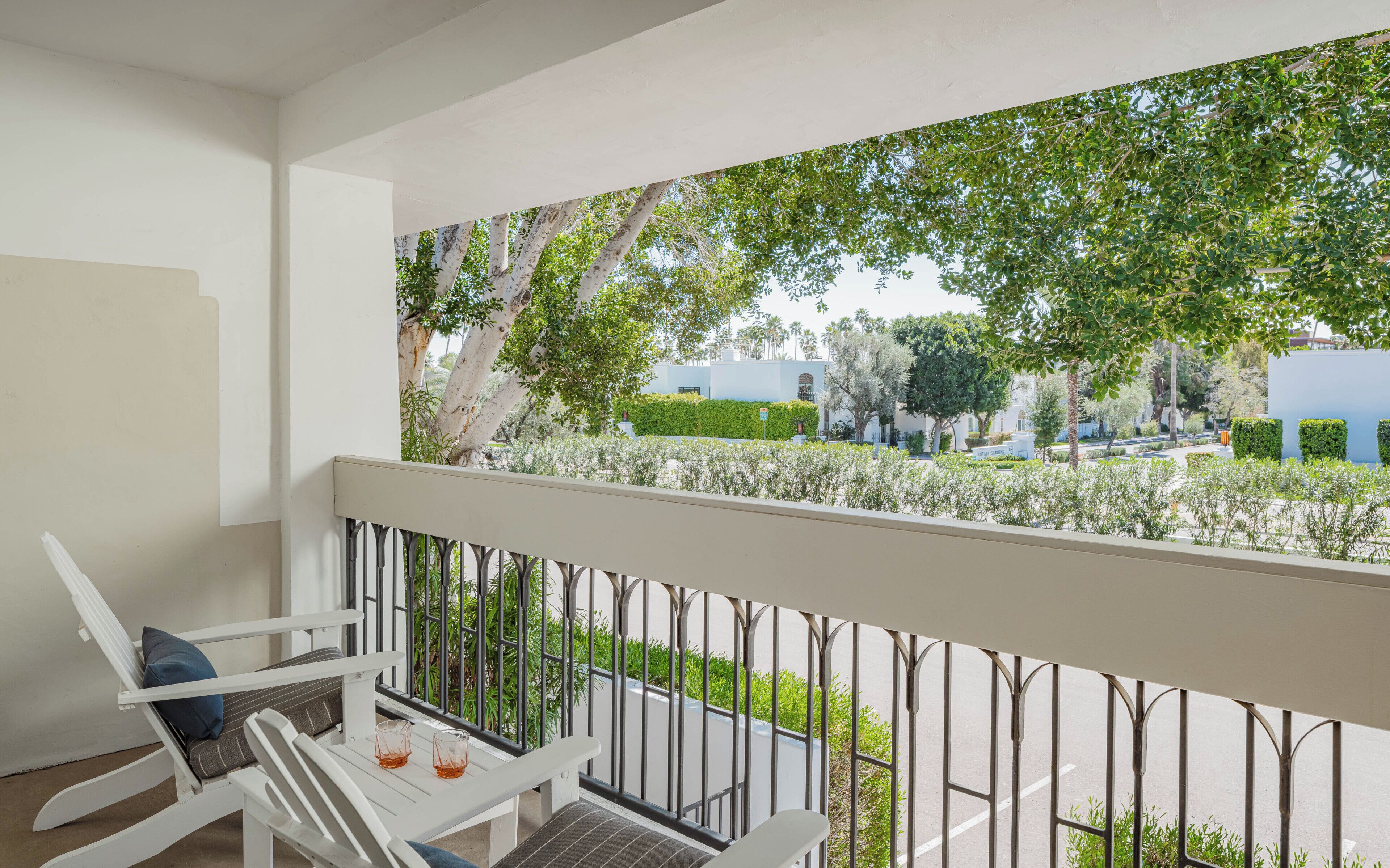 A balcony with a white railing, a white chair, a small table with two glasses, and a view of a green landscape with trees and buildings.