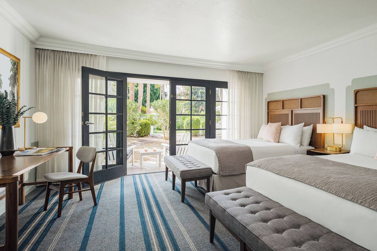 A hotel room with a large bed, a desk with a chair, a small table, and a bench. There is a door leading to an outdoor area with plants and a view of the outdoors.