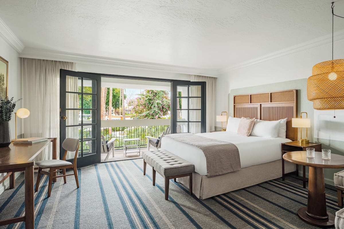 A hotel room with a bed, a desk, a chair, a round table, a bench, and a balcony with a view of greenery.