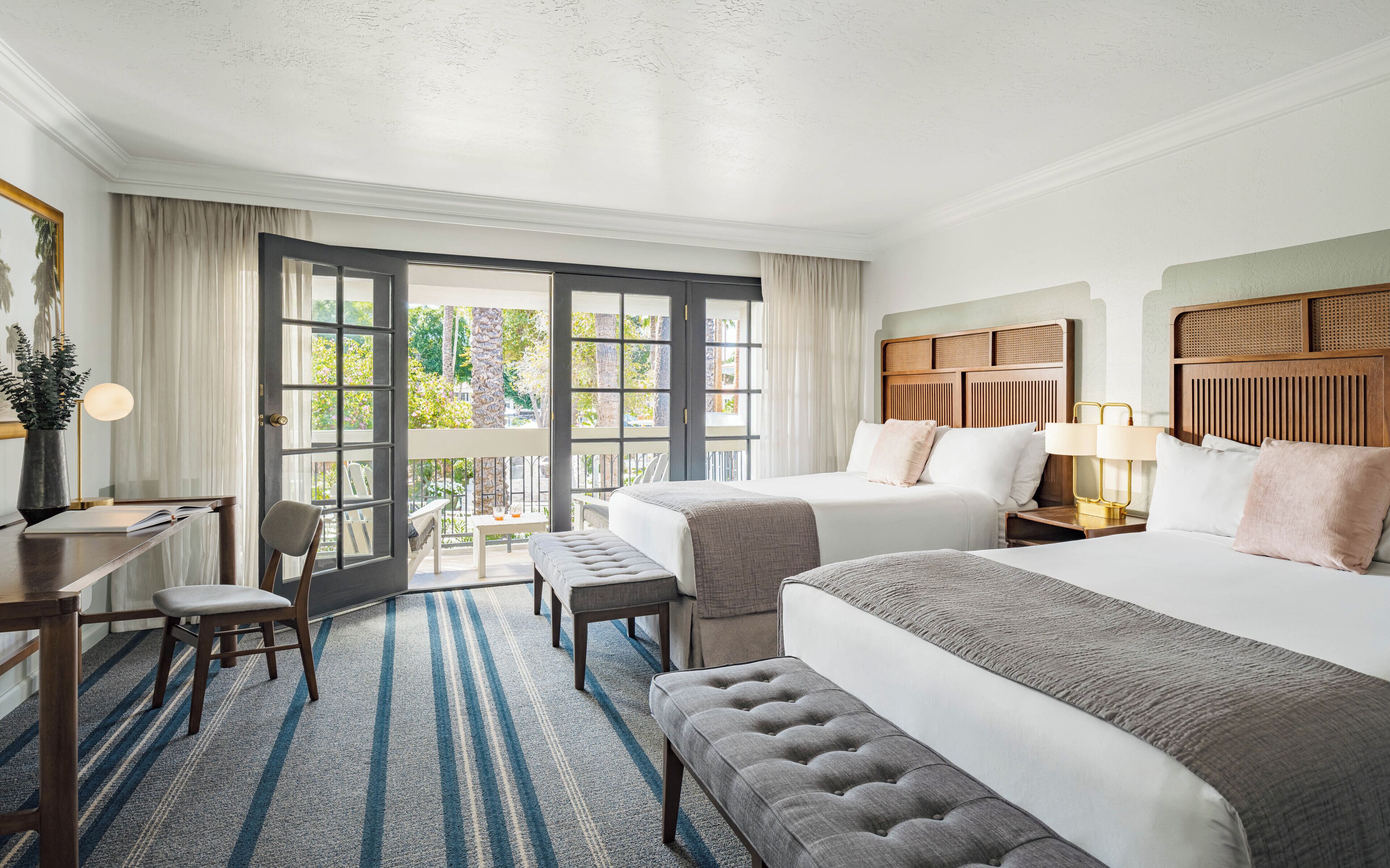 Ein Hotelzimmer mit zwei Betten, einem Schreibtisch, einem Sessel, einem kleinen Tisch und einem Balkon mit Blick auf Bäume.