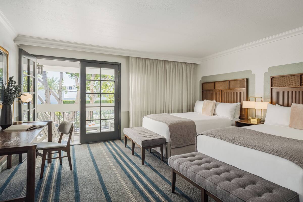 A hotel room with two beds, a desk, a chair, a bench, a large window with curtains, and a view of palm trees.