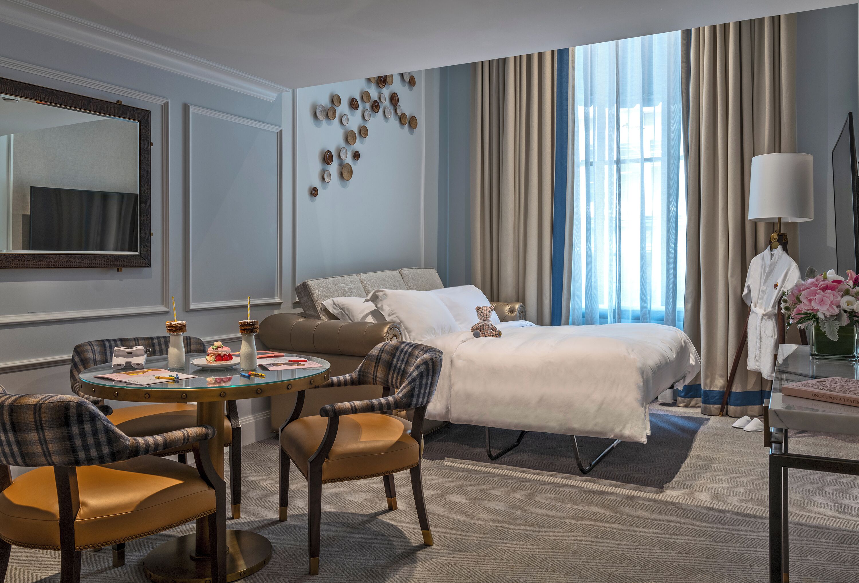 A hotel room with a bed, a round dining table with chairs, a sofa, a mirror, and a large window with curtains.