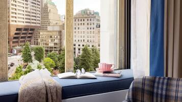 A window seat with a view of city buildings, a book, a pair of glasses, and a pink cup.