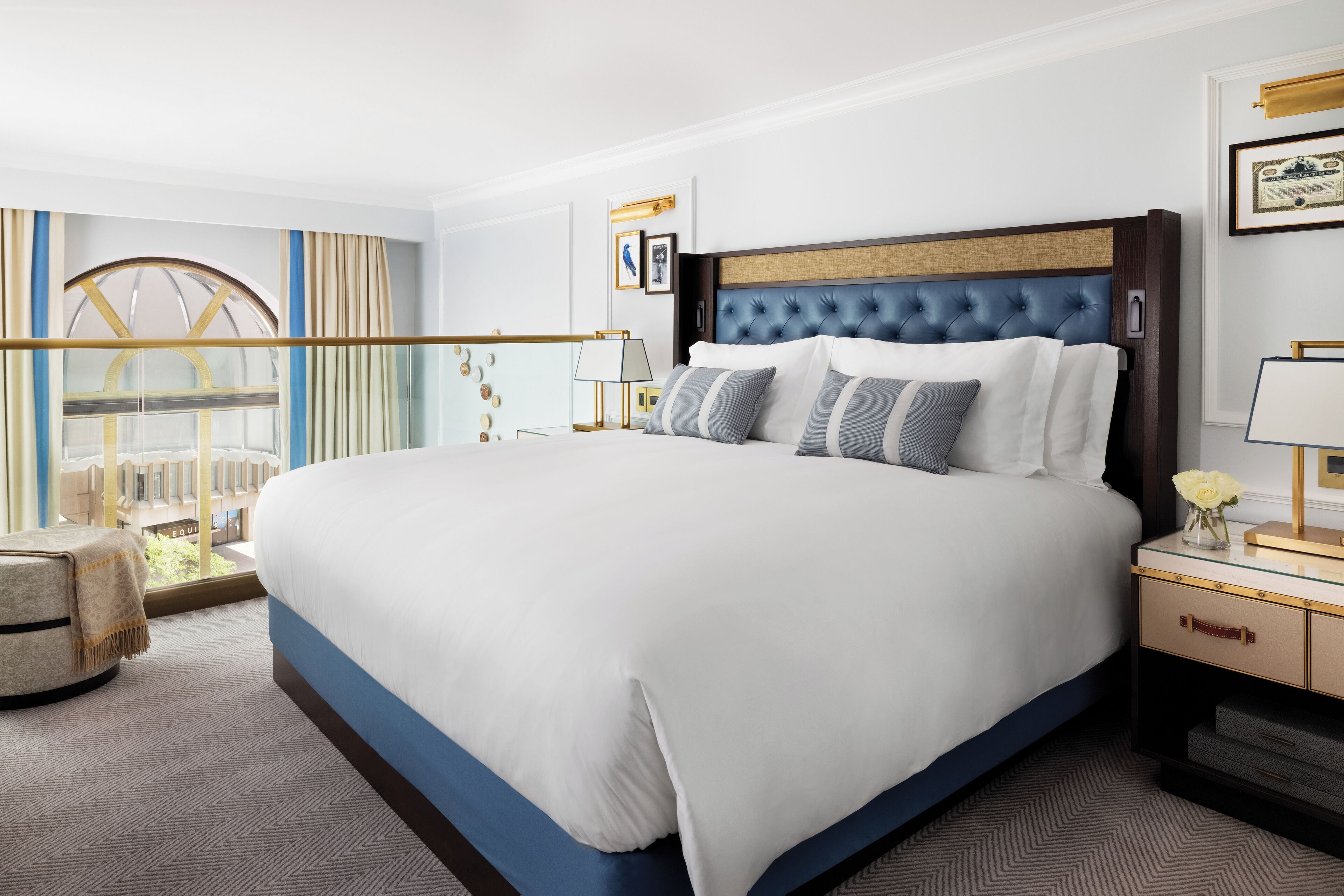 A hotel room with a large bed, a nightstand, a lamp, and a view of a building through a window.