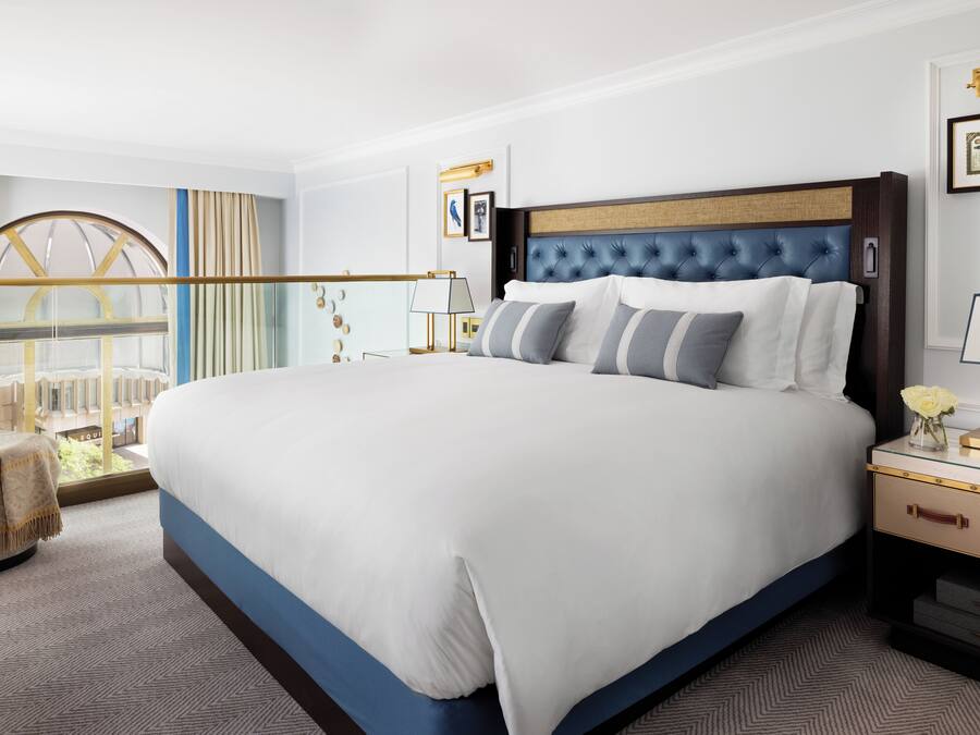 A hotel room with a large bed, a nightstand, a lamp, and a view of a building through a window.