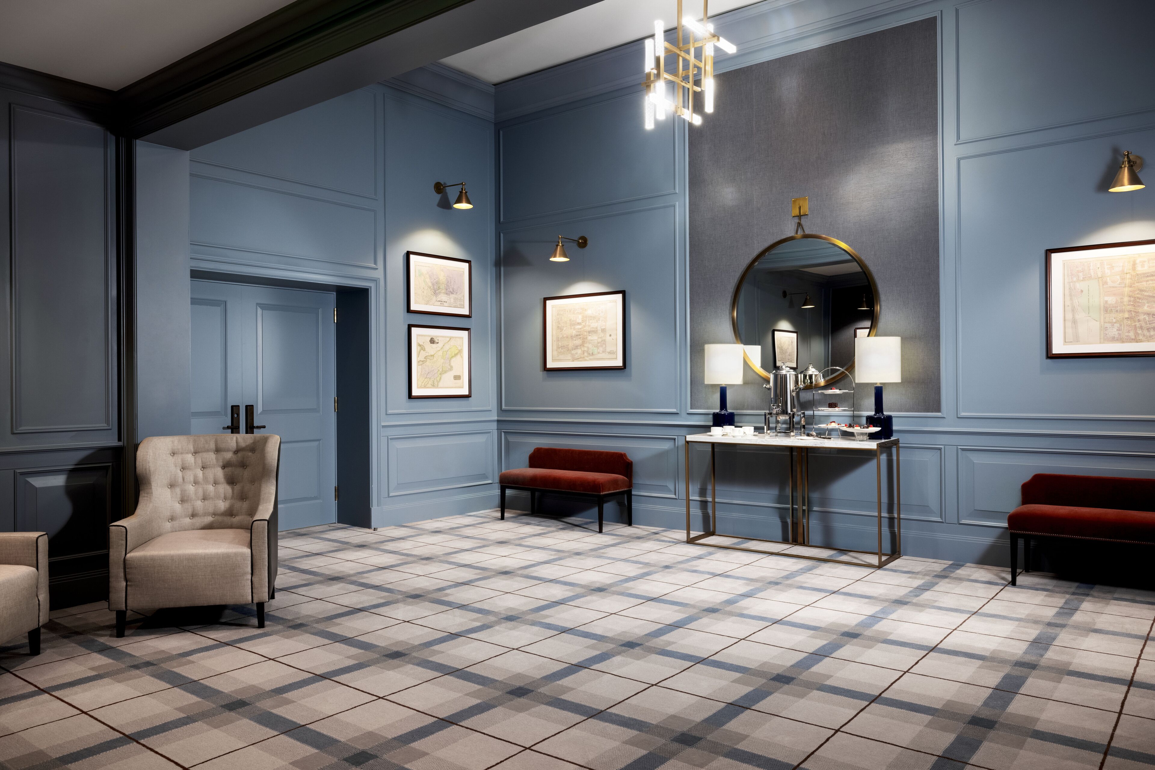 A room with a checkered floor, a tufted armchair, a red bench, a table with a mirror, and framed maps on the wall.