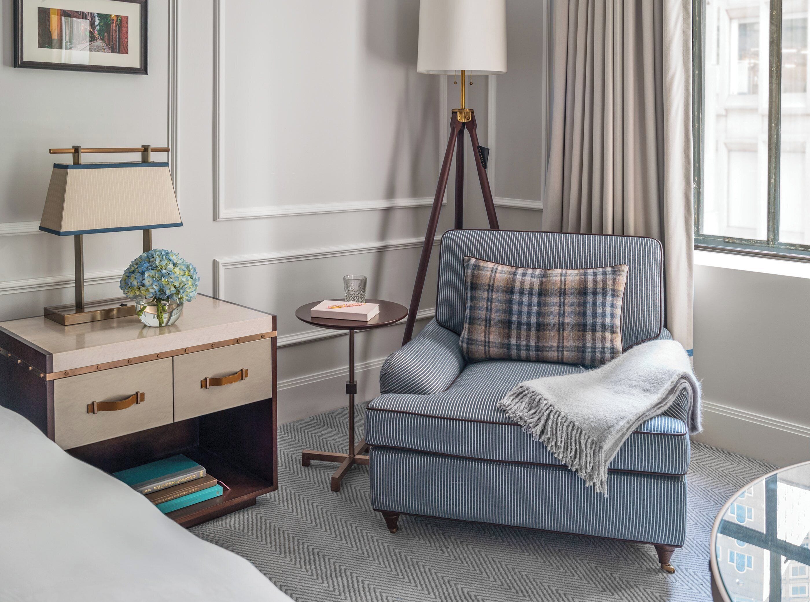 A hotel room with a bed, a bedside table with a lamp, a small round table with a glass, a striped armchair with a pillow, and a view of buildings through the window.