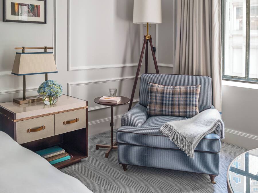 Habitación de hotel con cama, mesita de noche con lámpara, una pequeña mesa redonda con un vaso, sillón a rayas con un cojín y vista a edificios a través de la ventana.