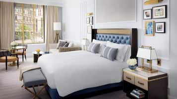 Habitación de hotel con una cama grande, una mesita de noche con lámpara, una silla y vistas a la ciudad a través de la ventana.