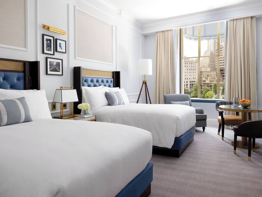 A hotel room with two beds, a desk with a chair, a lamp, and a view of the city through large windows.