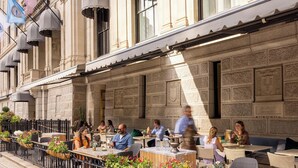 An outdoor cafe with tables and chairs, a stone facade, and flags. - The Langham, Boston (Boston)