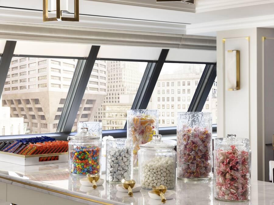 A candy display with various types of candies in clear jars, set on a marble counter with a city view through large windows.