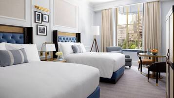 Habitación de hotel con dos camas, un escritorio con silla, una lámpara y vistas a la ciudad a través de amplios ventanales.