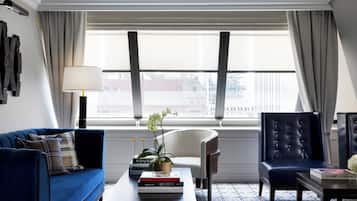 A modern hotel room with a blue sofa, a white armchair, a leather armchair, a coffee table with books and a plant, and a large window with curtains.