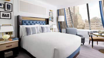 A modern hotel room with a large bed, a seating area with a table, and a view of the cityscape through large windows.