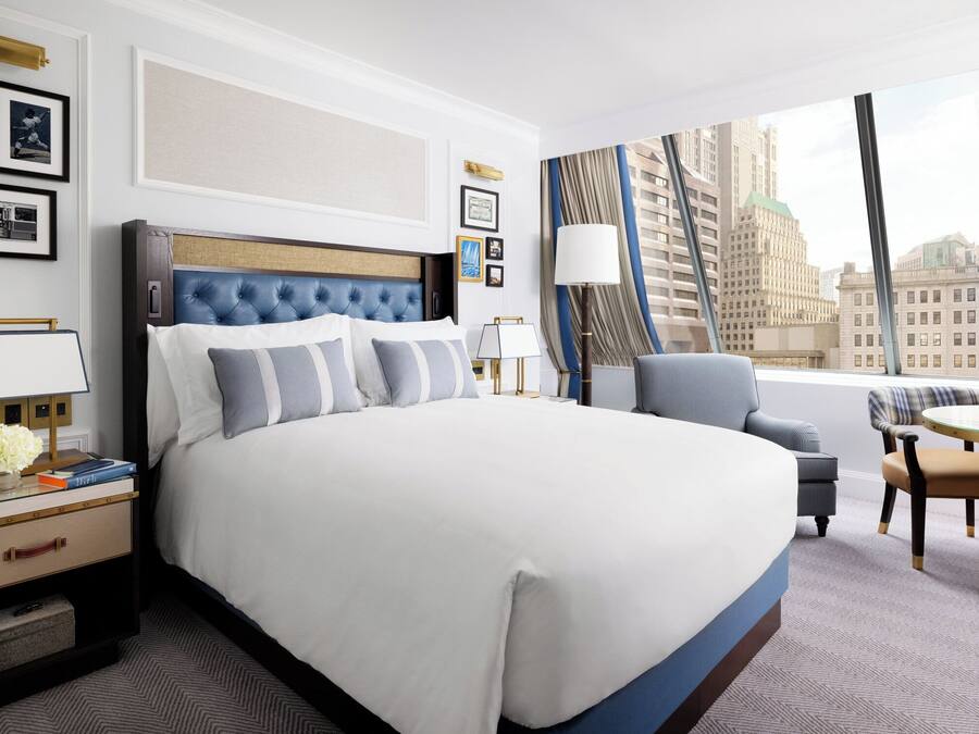 A modern hotel room with a large bed, a seating area with a table, and a view of the cityscape through large windows.